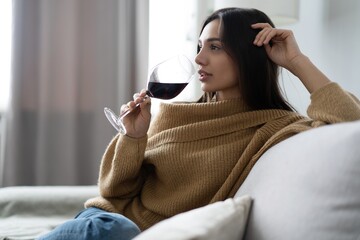 Pretty, attractive, stylish young woman sitting on couch having raised glass with red wine, examine, taste beverage