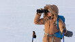 © junky_jess - Woman in yellow hooded down jacket with a backpack and ski poles looks through binoculars standing on a snowy plain in winter, waist up, selected focus.