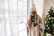 © nikkimeel - Young caucasian woman standing near window and using mobile phone, drinking tea in winter morning.