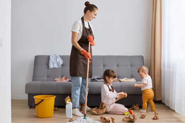 Smiling optimistic woman with bun hairstyle wearing white t shirt, brown apron and jeans, cleaning house, posing with her little children, young housewife washing floor and looking after kids.