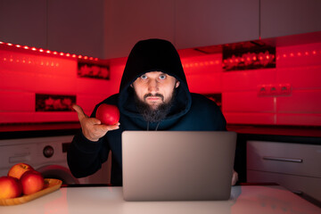 a hacker in a black hoodie with a hood on his head is sitting at a laptop holding a red apple in front of him