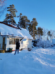 A young man in a warm jacket shovels the snow with a shovel, making the road along a country house