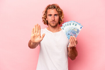 Wall Mural - Young caucasian man holding banknotes isolated on pink background standing with outstretched hand showing stop sign, preventing you.