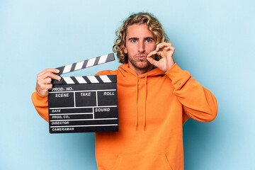 Wall Mural - Young caucasian man holding a clapperboard isolated on blue background with fingers on lips keeping a secret.