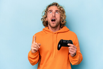 Wall Mural - Young caucasian man playing with a video game controller isolated on blue background pointing upside with opened mouth.