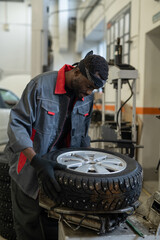 Wall Mural - Vertical portrait of young black mechanic inspecting car tires while working in auto repair shop