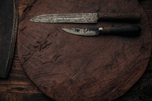 Two Vintage Knives On An Old Wooden Round Cutting Board Near A Cleaver Blade