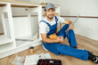 © Kostiantyn - Smiling handyman in overalls holding a part of kitchen cabinet while installing furniture, sitting on the floor