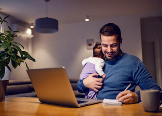 A smiling businessman working online job from home and holding his baby girl in his arms.