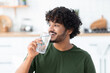 © kucherav - Happy young Indian man standing at home in the kitchen drinking clean water from a glass, looks looking away and smiles friendly. Healthy lifestyle concept