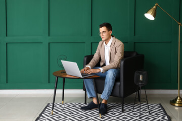 Wall Mural - Young man with eyeglasses using laptop in armchair near green wall