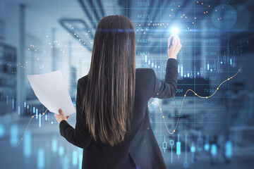 Canvas Print - Back view of businesswoman with accounting document pointing at abstract glowing big data forex candlestick chart on blurry office interior background. 