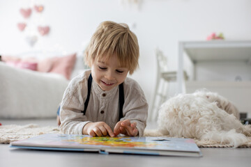 Poster - Cute blond preschool child, blond boy with pet maltese dog, reading book at home with mother