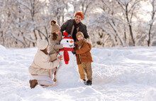 Happy Children Sculpting Funny Snowman Together With Parents In Winter Snow-covered Park