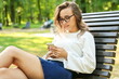 © Petr Smagin - young woman holding phone and sitting on the bench in the summer park. girl reading sms message on her phone outdoors close up.