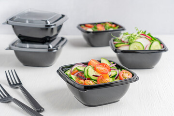 Wall Mural - Variety of vegetarian healthy vegetable salads made of sliced cucumber, tomatoes and radish served in black plastic bowl and fork on white wooden background. Food delivery concept and healthy eating
