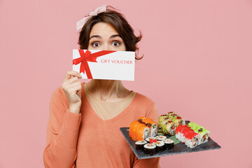 Wall Mural - Young woman in sweater hold in hand makizushi sushi roll served on black plate traditional japanese food cover mouth with gift certificate coupon voucher card isolated on plain pastel pink background