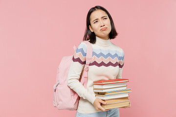 Wall Mural - Disappointed sad teen student girl of Asian ethnicity wearing sweater backpack hold pile of books look camera isolated on pastel plain light pink background Education in university college concept