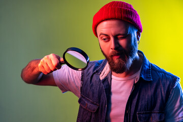 Hipster man looking through magnifying glass, spying, finding out something, exploring and inspecting, wearing beanie hat and denim vest. Indoor studio shot isolated on colorful neon light background.