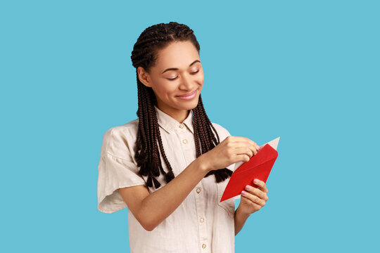 Portrait of smiling woman with black dreadlocks standing open red envelope with congratulations, reading romantic letter, wearing white shirt. Indoor studio shot isolated on blue background.