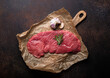 © somegirl - Beef lean raw fillet steak on baking paper with rosemary, garlic and spices on dark brown rustic concrete background with knife from above flat lay, diet beef meat steak ready to be cooked, copy space