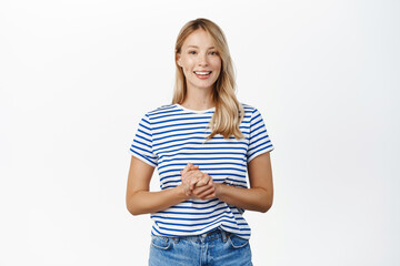 Wall Mural - People concept. Stylish modern girl, 25 years old, standing in assistant helpful pose, ready to offer help, listening to customer and smiling, standing over white background