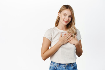 Wall Mural - People and faces concept. Young smiling woman holding hands on heart and looking grateful, thank you gesture, feeling tenderness, standing over white background