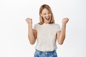 Wall Mural - Enthusiastic young woman winning on mobile phone, celebrating and cheering, shouting with joy, standing over white background