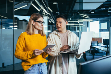 Brainstrom business team is responsible for solving the tasks, employees colleagues Asian man and woman work together in a modern office, look at the screen laptops, a joint IT project