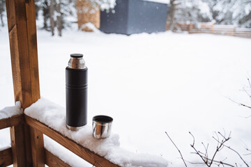 black matte thermos and it's metal mug with hot drink on window sill of the outdoor veranda. Hiking and picnic staff. Drinking warm tea or coffee in the winter frost.