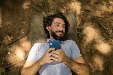 View of young man using a smartphone at day time lying down on a bench at a park. High quality photo