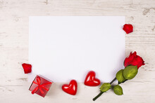 A Red Rose, Two Decorative Hearts And A Gift Box Lie On A Table With A White Sheet Of Paper. Mockup For Writing Text Congratulations On Valentine's Day Or Declaration Of Love, Or Wedding