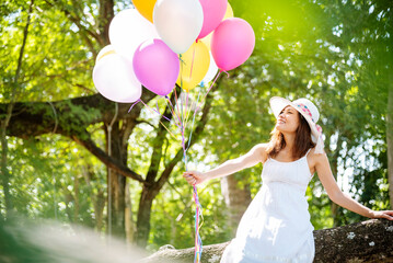 Cheerful beauty woman holding balloons relax sitting under big tree in green park with happiness. Woman Hands holding vibrant air balloons play on birthday party happy time summer on sunshine outdoor