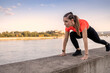 Young happy focused fitness girl in black yoga pants and orange short shirt work out and stretch her body on short concrete wall near river during the day.