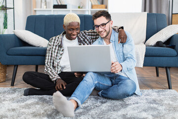 Wall Mural - Happy multiracial couple of two gays sitting on floor at living room and using modern laptop. People in love scrolling social networks during free time.