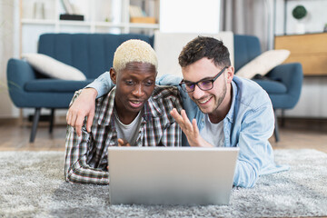 Wall Mural - Two multiracial gays in casual clothes resting on soft carpet and using wireless laptop. Happy loving people watching funny videos during free time at home.