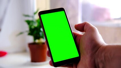 A hand holds a mobile phone with a green screen in a living room.