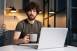 © dikushin - Portrait of confused curly young freelancer male holding in hand cup with morning coffee sitting at table with laptop computer, looking at camera, in kitchen with modern interior, remote home office.