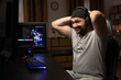 © ABCreative - A man playing professionally on a computer with a headset puts his hands behind his head in anger, disappointment, lost a round in a video game, a middle-aged guy in a dark room