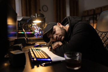 Tired sleepy overworked man in cap and sweatshirt works at night in dark room, sleeps in front of computer with head on desk on notes, next to mug of coffee lighted lamp