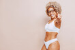Cheerful beautiful afro latin american woman in white underwear and eyeglasses pointing a finger at camera, isolated on beige background at studio. Real body, natural beauty.
