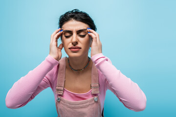 young woman with closed eyes touching head while suffering from migraine isolated on blue