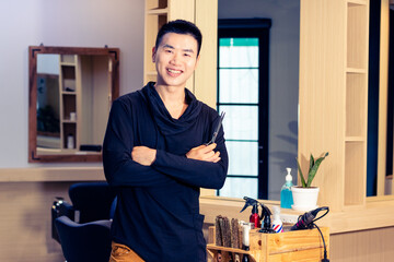 Hairdresser hold scissors, standing and smiling in salon. Young Asian man LGBT hairstylist ready to welcome and service customer in barbershop. Haircut and hair coloring concept.