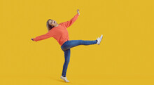 Funny Carefree Young Woman In Comfortable Casual Wear Having Fun And Fooling Around. Happy Pretty Teenage Girl In Orange Sweatshirt And Blue Jeans Dancing In Studio With Vibrant Yellow Background
