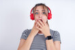 © Jihan - Shocked young Arab woman wearing striped t-shirt over white background stares fearful at camera keeps mouth widely opened wears wireless stereo headphones on ears