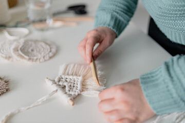 Woman working out last details of an almost-finished macrame piece, combing ropes inside a home room. Close up. Natural cotton threads and wooden beads. Female hobby. Wall hanging decor.