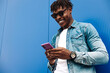 happy curly african american man wearing sunglasses using mobile phone and smiling against blue wall background