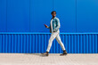 © Shopping King Louie - stylish African American man in full growth, uses a smartphone and walks past a blue wall while going to work