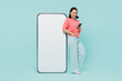 © ViDi Studio - Full body young smiling happy woman of Asian ethnicity 20s in pink sweater stand near big mobile cell phone with blank screen workspace area chatting isolated on pastel plain light blue background
