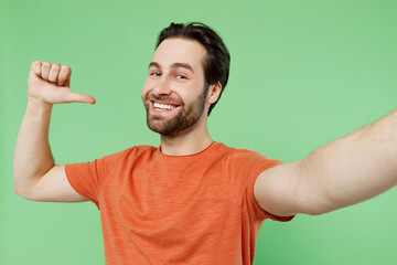 Close up young exultant happy fun cool man 20s in casual orange t-shirt doing selfie shot pov on mobile phone point finger on himself isolated on plain pastel light green background studio portrait.